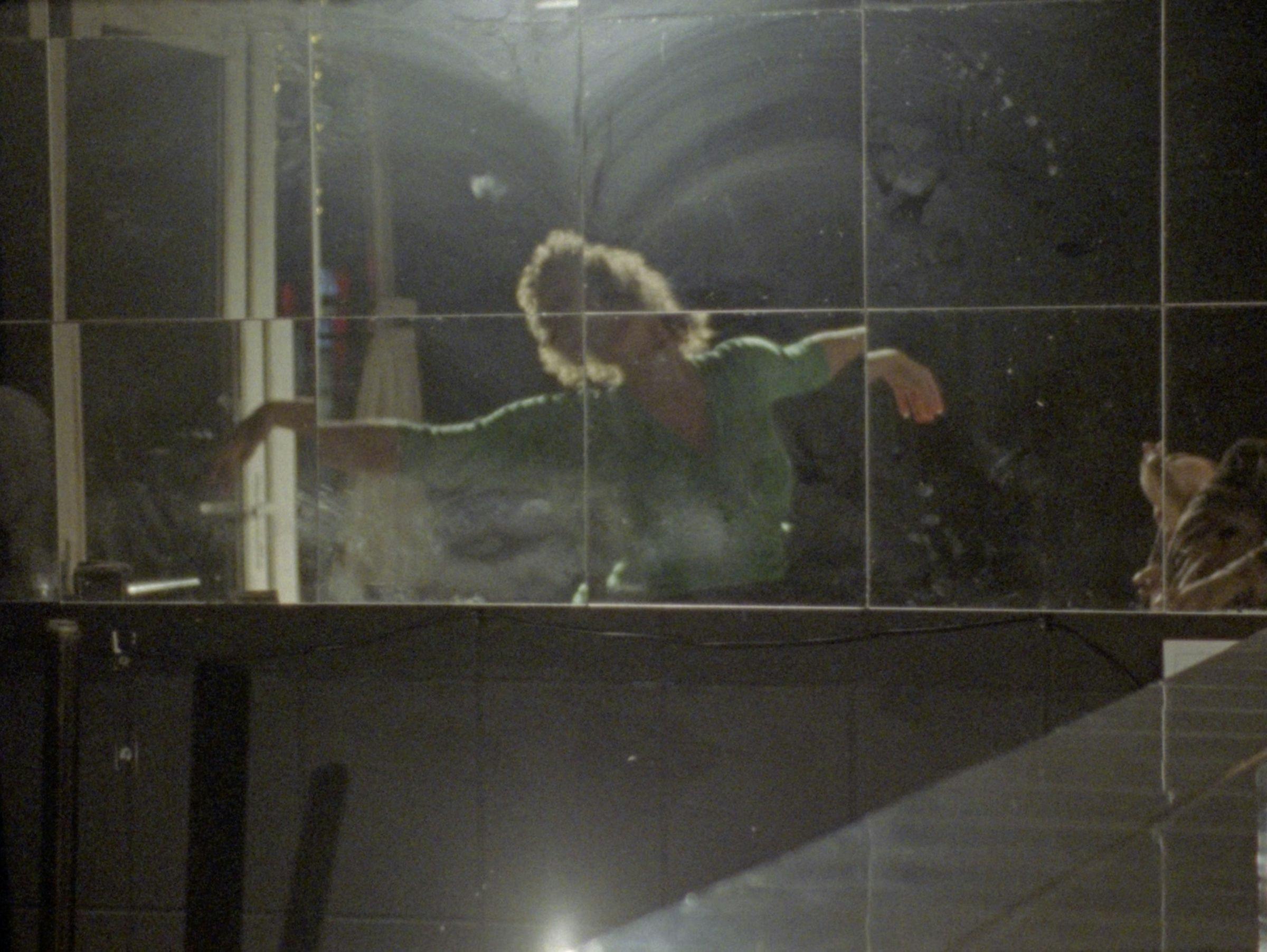 a dancer wearing a green leotard with out streched arms is reflected in a mirrored wall in bar