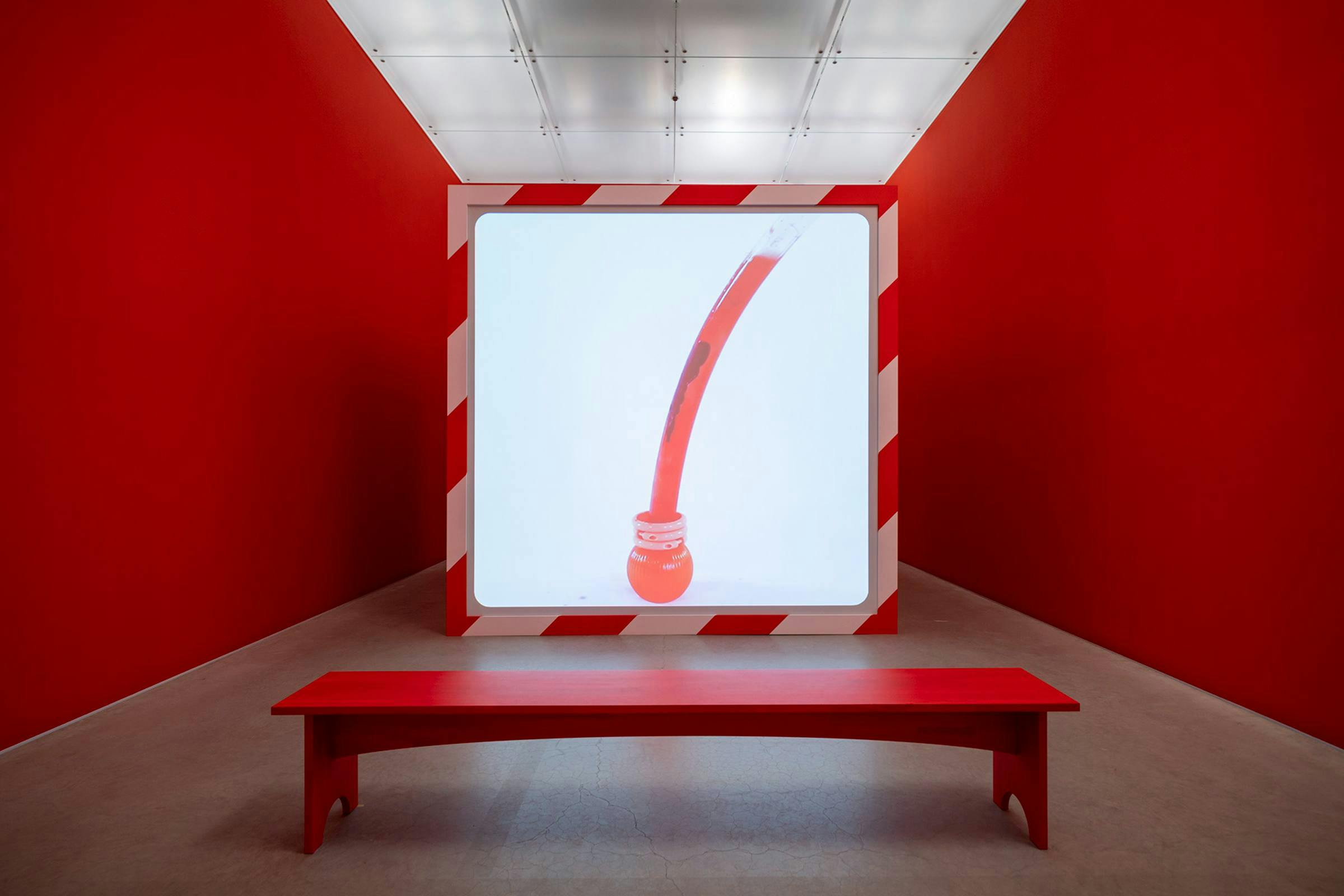 Red painted gallery space with red bench in front of a large screen with a white background and red object projected on top.