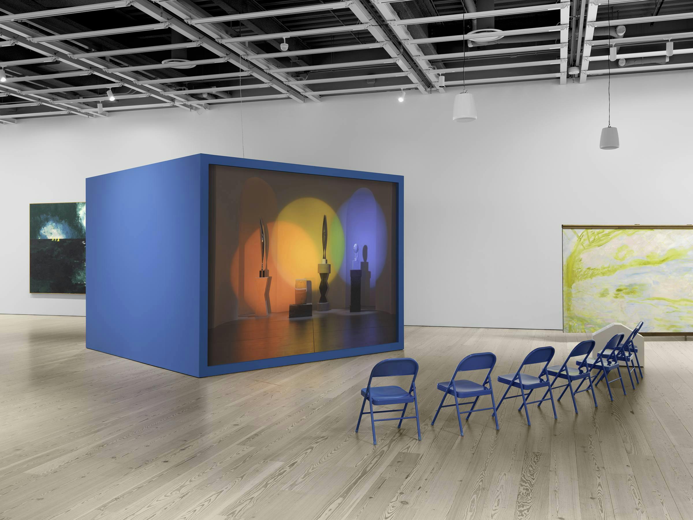 an art gallery with wooden floors and white walls. A video art work projected on to a blue freestanding box. 8 blue chairs are in front of the projection for the audience to sit on. 