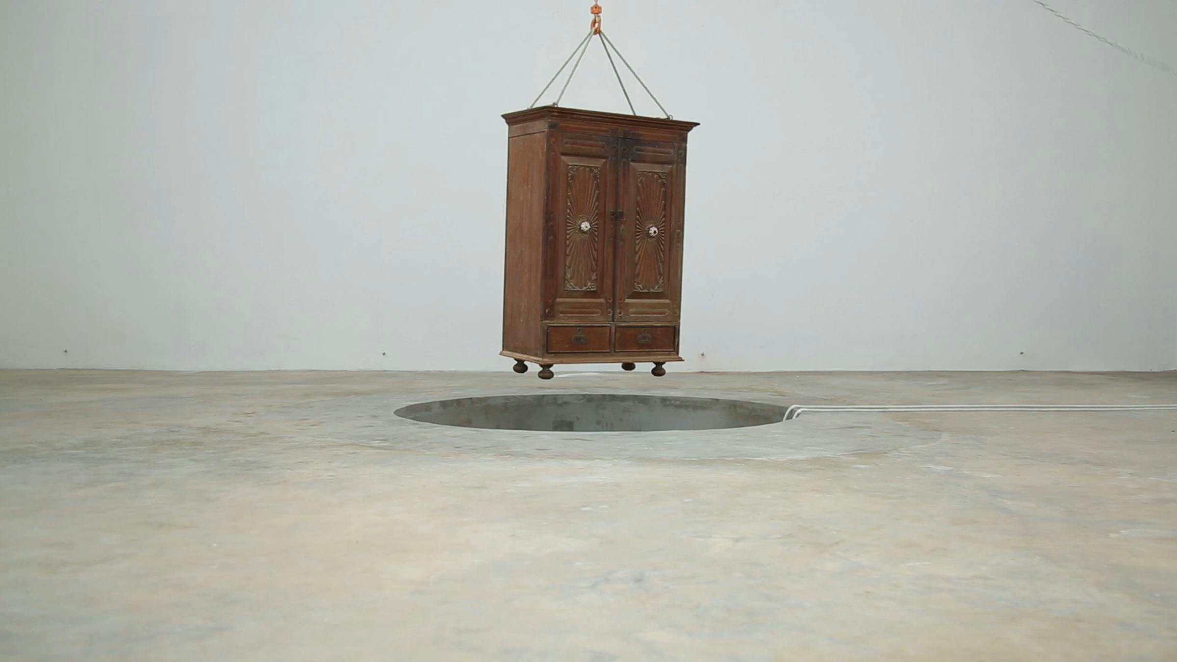 A wooden cupboard is held by a winch above a hole in a concrete floor. The room is painted 