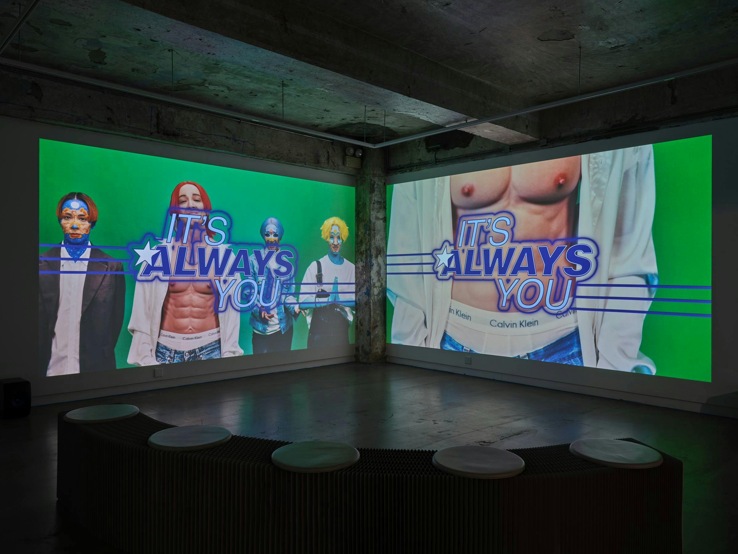 a gallery space with 2 projections that meet in a corner. There is seating in front of the projections. The space is industrial.