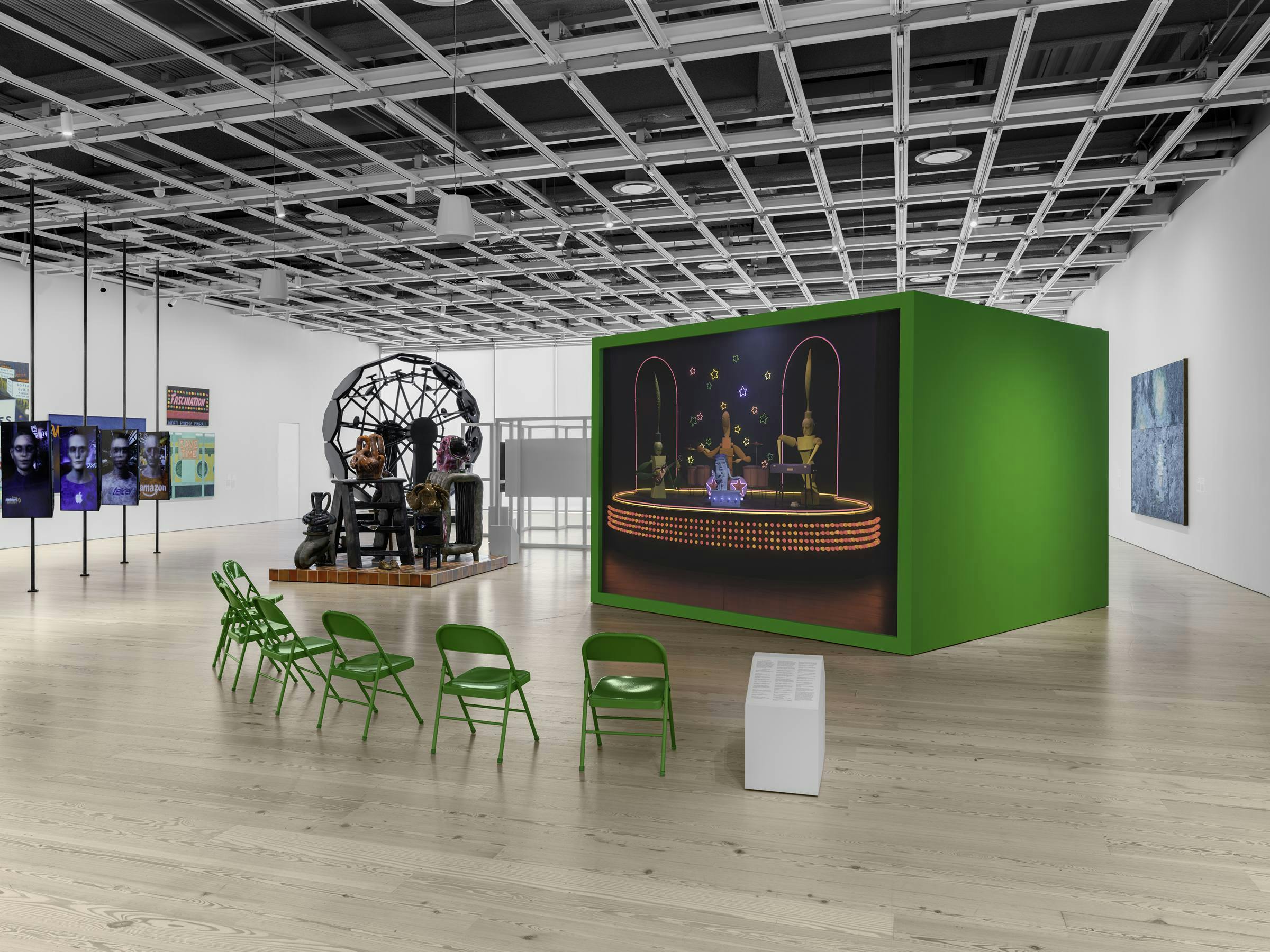 an art gallery with wooden floors and white walls. A video art work projected on to an green freestanding box. 8 green chairs are in front of the projection for the audience to sit on. 