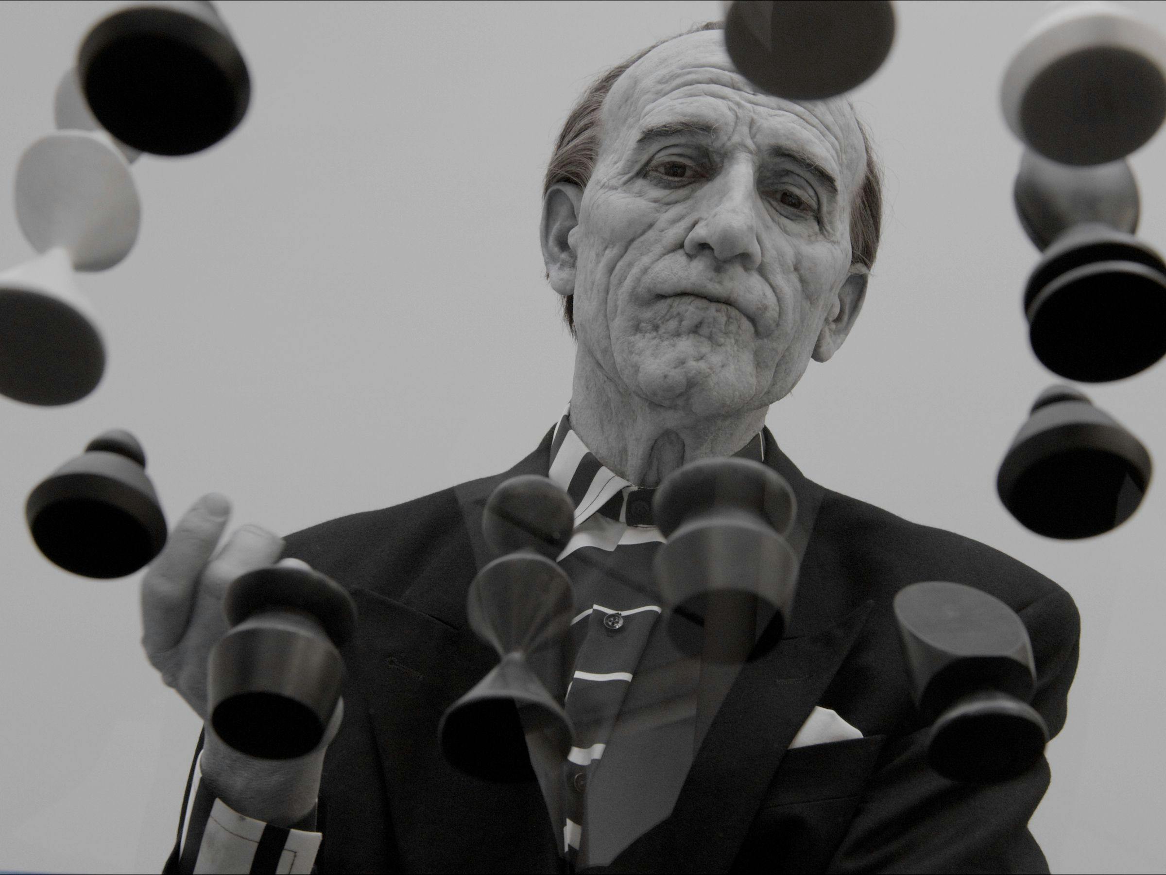 a black and white image of an elderly man wearing a black suit is playing chess. We can see him from underneath and through a glass table.