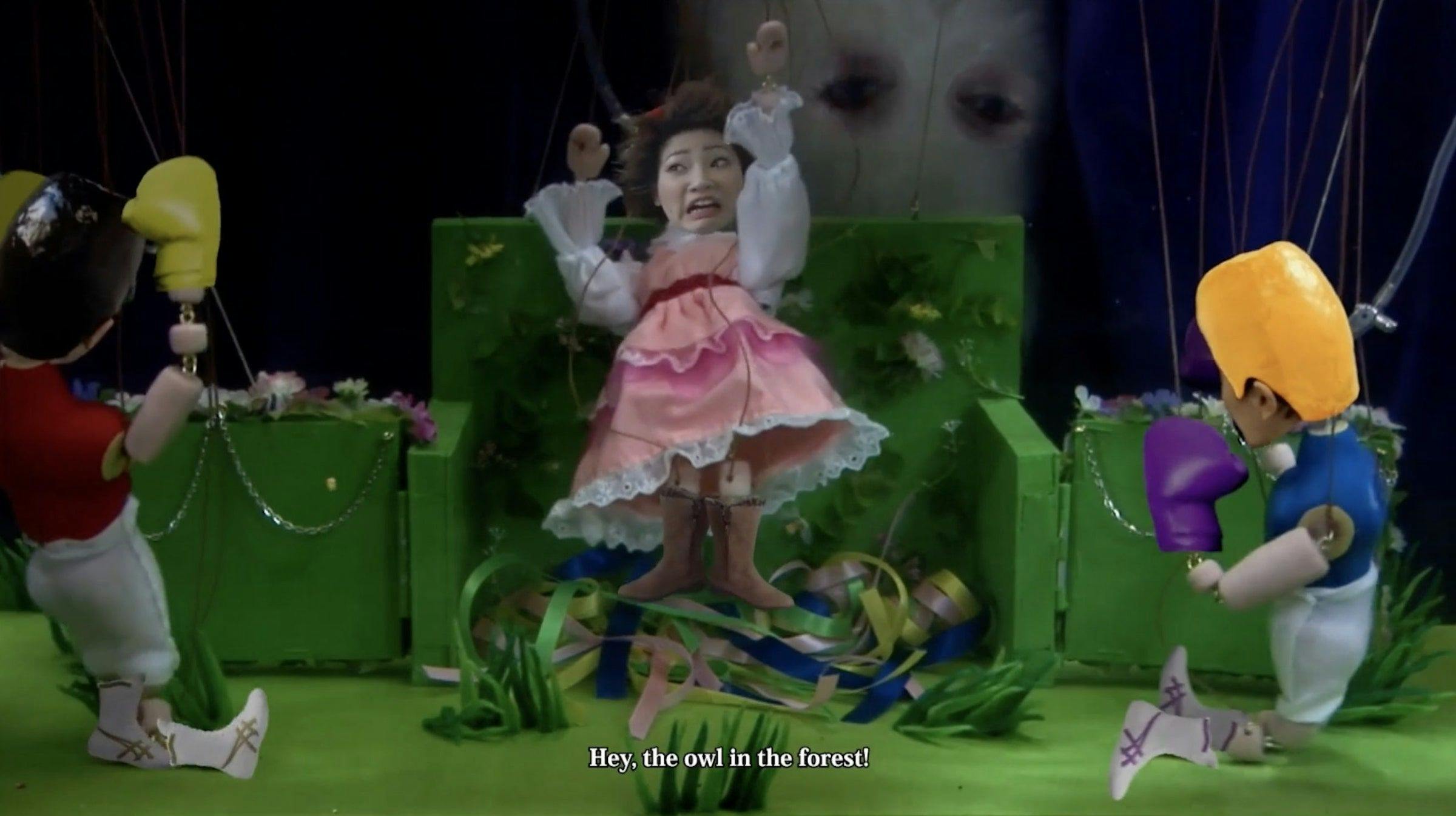a puppet has her arms up in the air in fright. She is wearing a pink dress. 2 other puppets with yellow helmets and boxing gloves face her in confrontation.